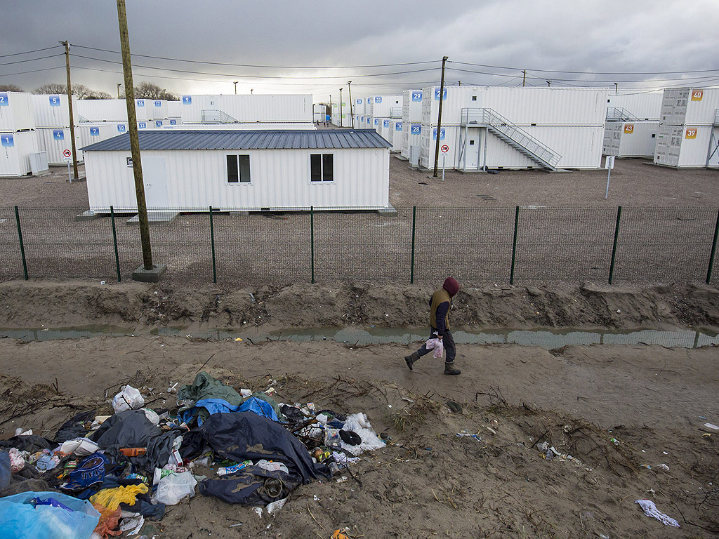 Мигрант се движи между палатките и бараките на импровизирания лагер, наречен Джунглата и нов лагер от контейнери, подготвени да се използват като жилища в Кале, Франция. Имигрантите, опитващи се дастигнат до Великобритания са между 4000 и 7000 в Кале
