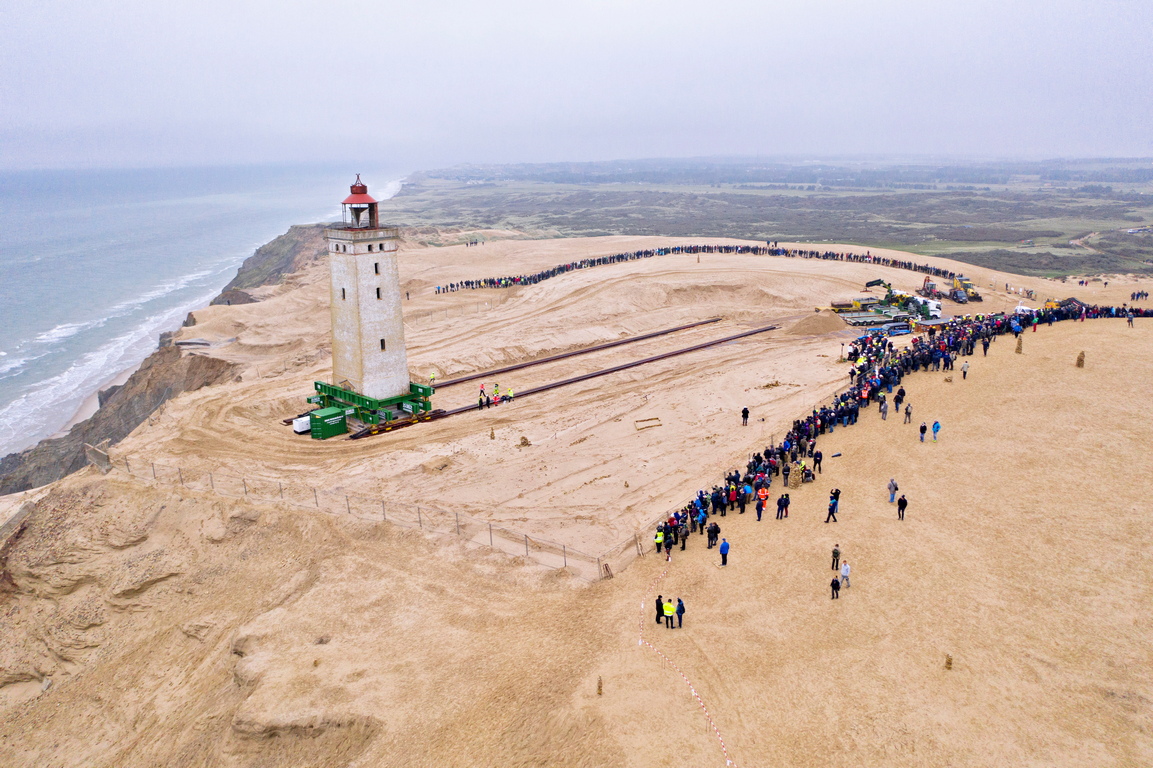 Работници и зрители следят преместването на фара Рубиерг Кнуде в Ютланд, Дания. 120-годишният фар е поставен на платформа и релси, за да го позиционират на около 80 метра от Северно море, тъй като е застрашен от крайбрежна ерозия и изместващи се пясъци.